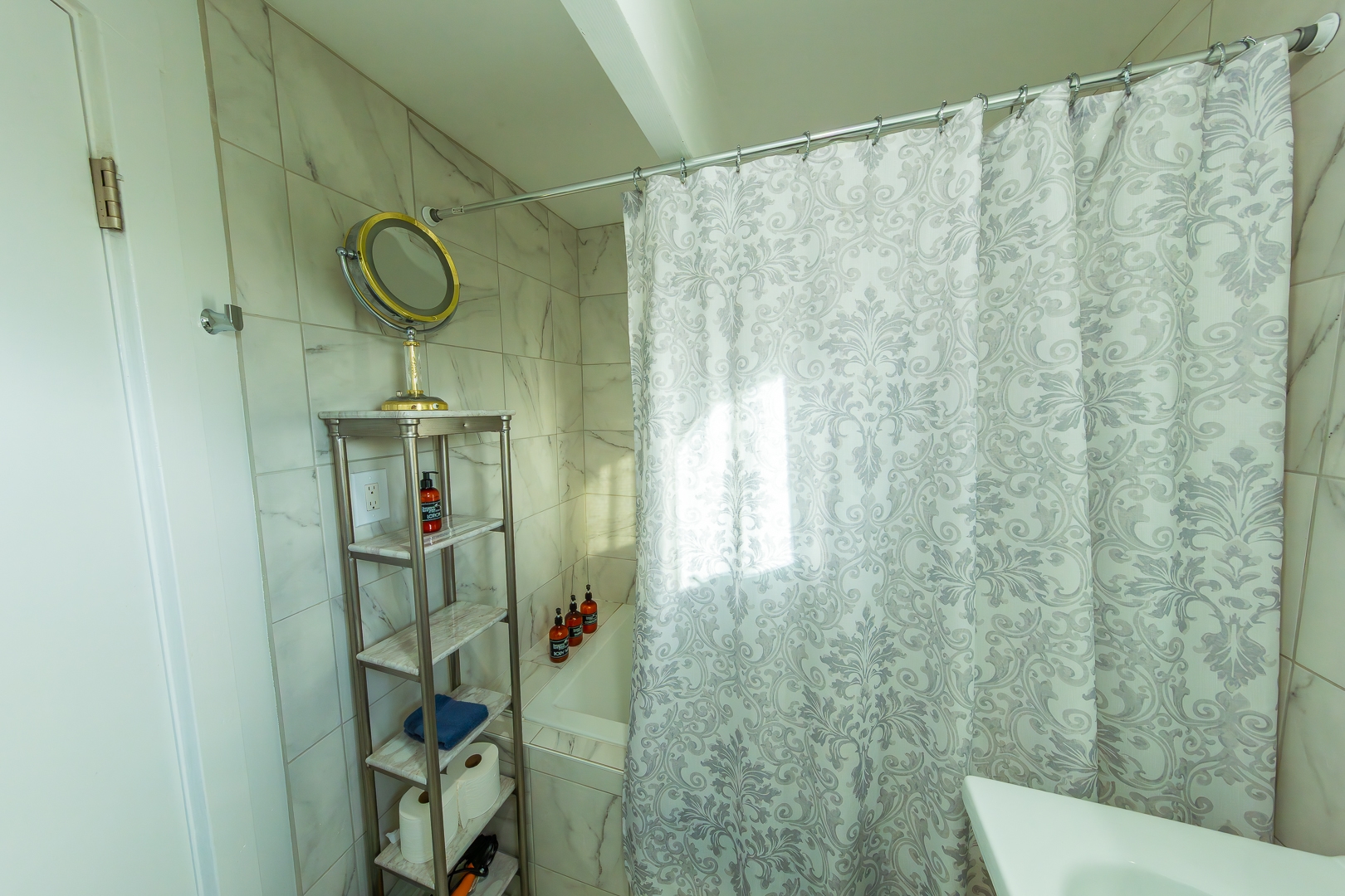 An elegantly tiled bathroom featuring a soaking tub/shower enclosed by a grey and white patterned shower curtain, complemented by marble-style wall tiles and a silver shelving unit holding toiletries and a vanity mirror.