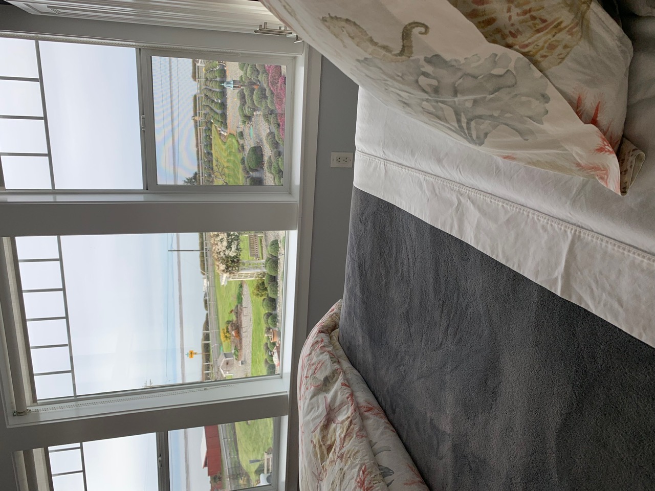 A view from the foot of a bed looking through large, white-framed windows toward a landscaped garden with rolling green lawns and a white garden arbor.