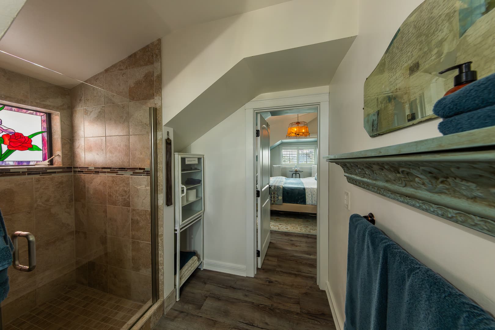 An updated, earth-toned tiled walk-in shower with a glass door, featuring a colorful red flower stained-glass window that casts a warm glow into the bathroom.