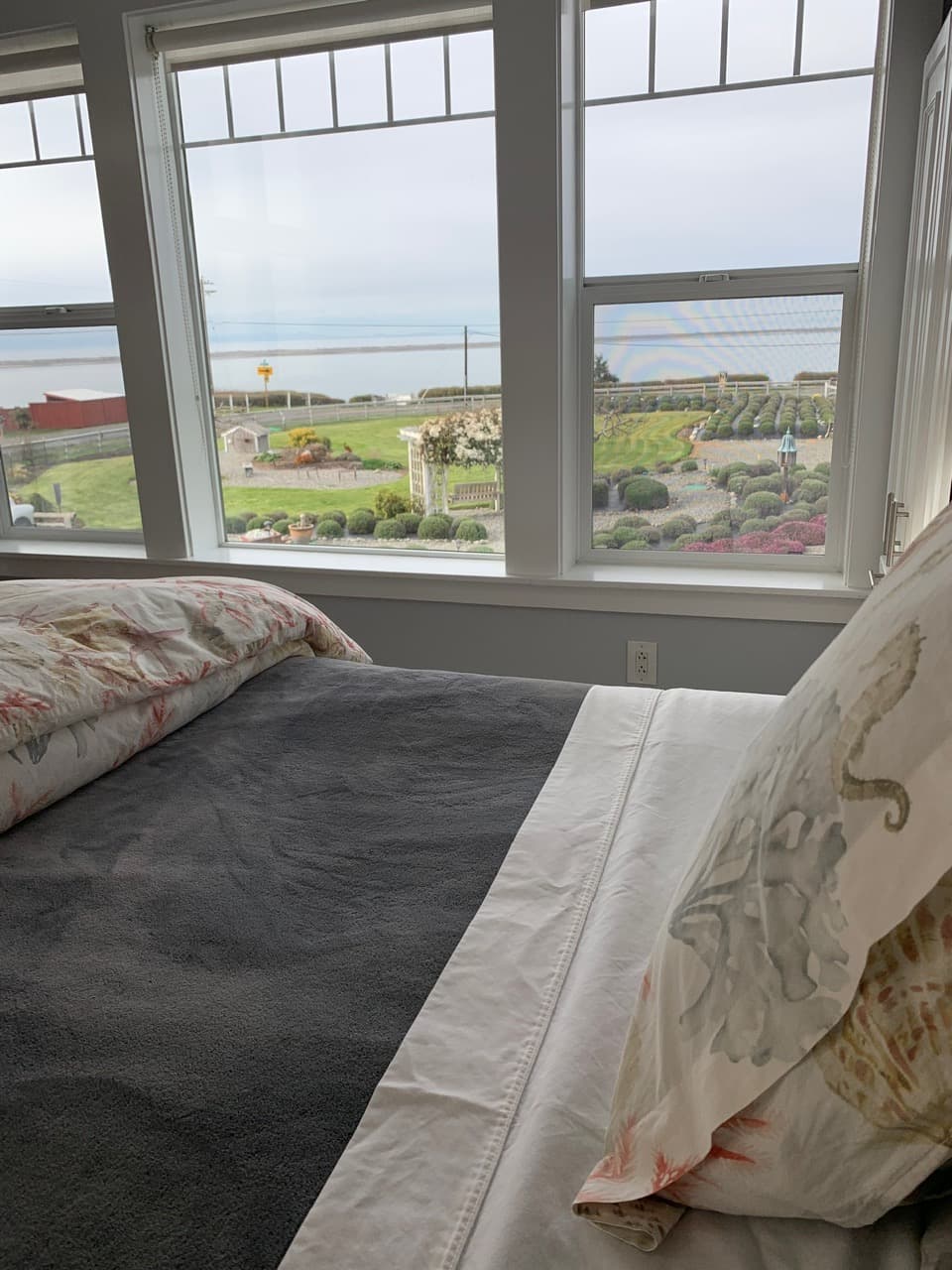 A view from the foot of a bed looking through large, white-framed windows toward a landscaped garden with rolling green lawns and a white garden arbor.
