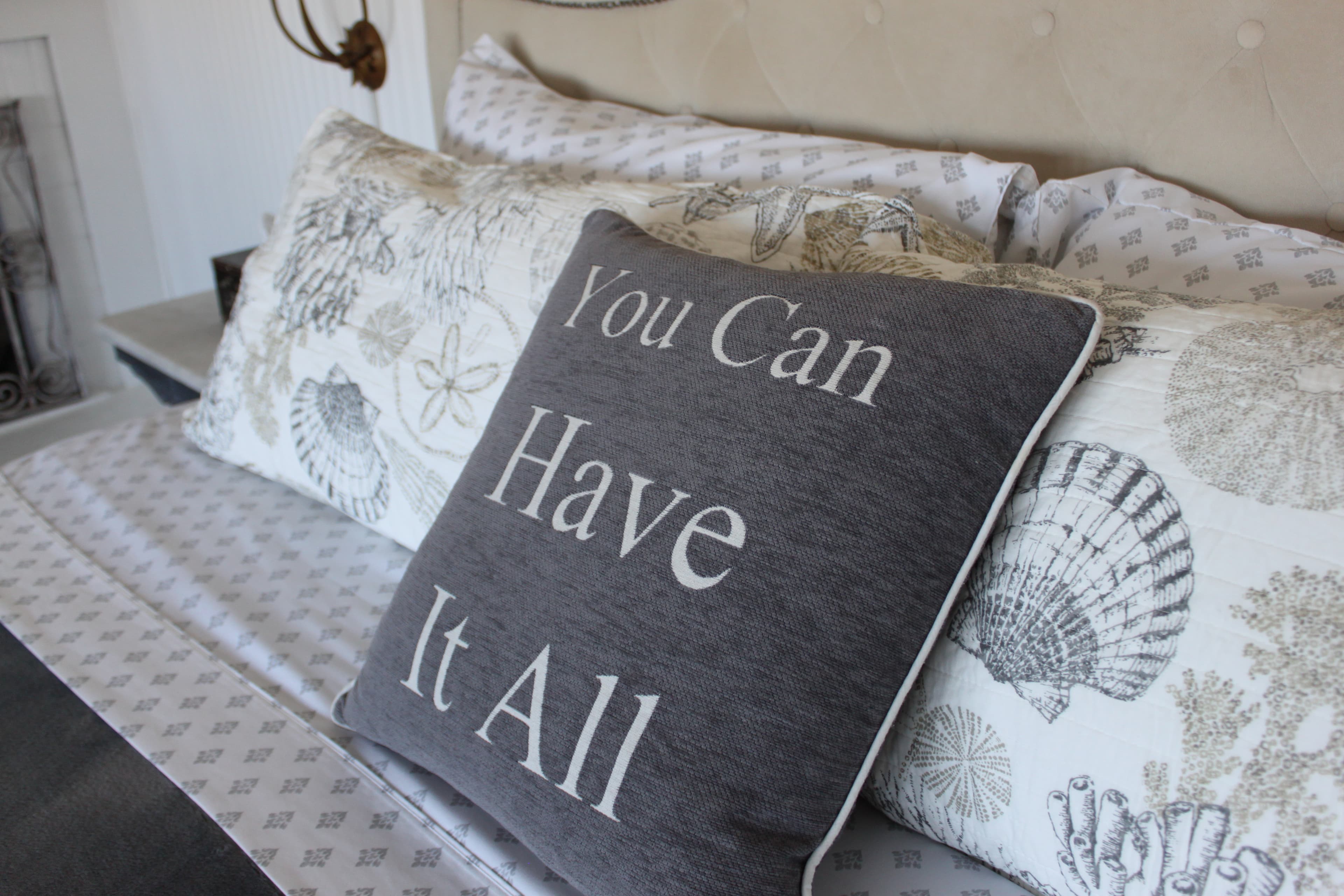 A close-up of a bed featuring decorative pillows with coastal seashell patterns and a dark grey accent pillow with the words "You Can Have It All" embroidered in white.