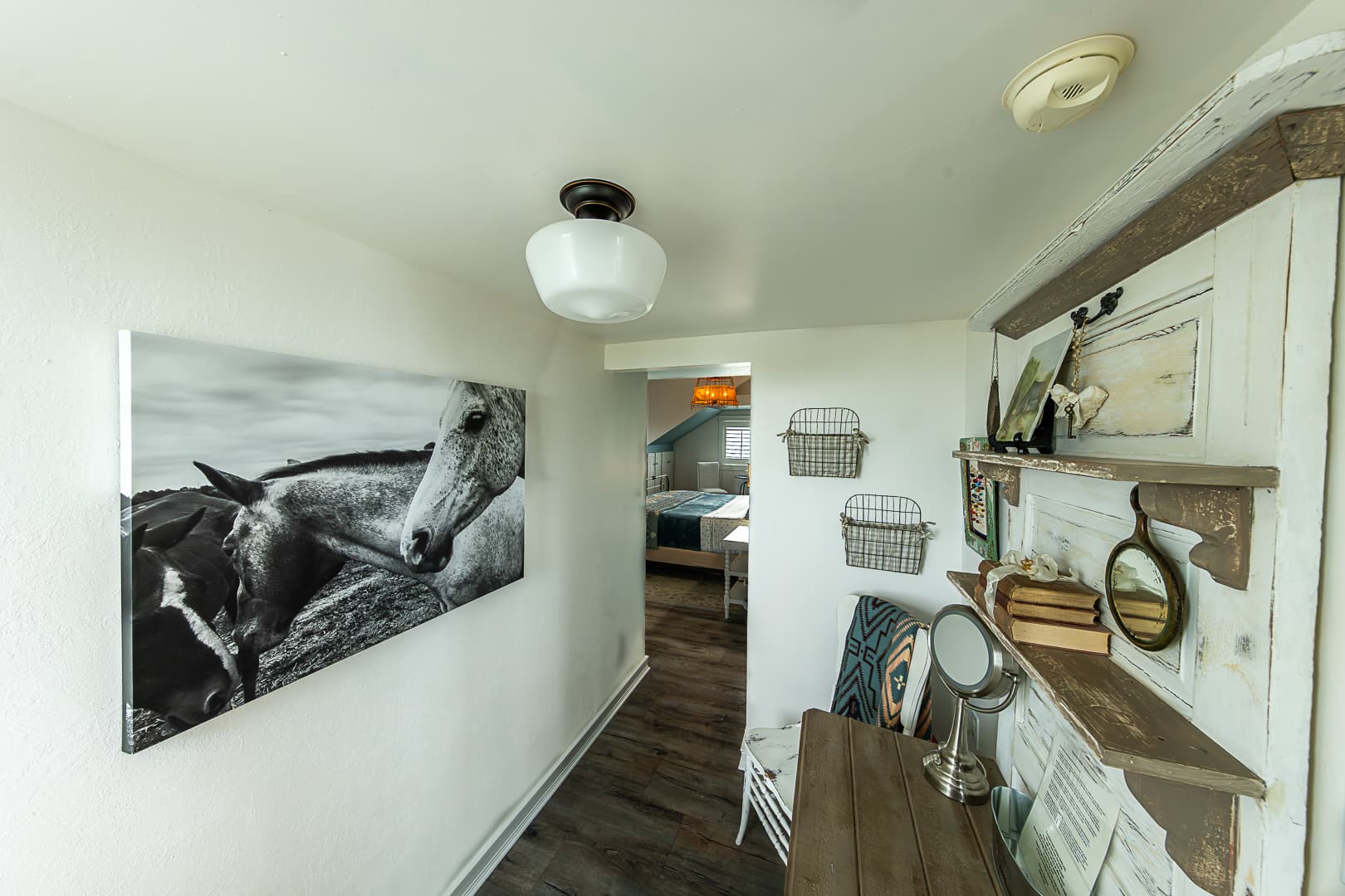 A narrow hallway featuring a large black-and-white canvas of a horse on the left wall and a rustic wooden shelf with vintage books and mirrors on the right, leading into a bright bedroom suite in the background.