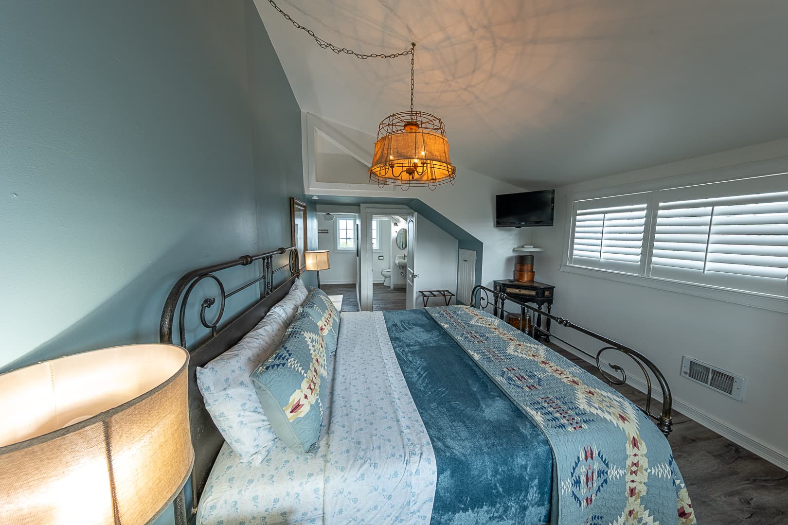 A wide view of the Garden View Suite featuring a king-size bed with light blue patterned linens, an ornate wrought-iron headboard, and a large orange-fringed pendant light illuminating the sloped ceiling.