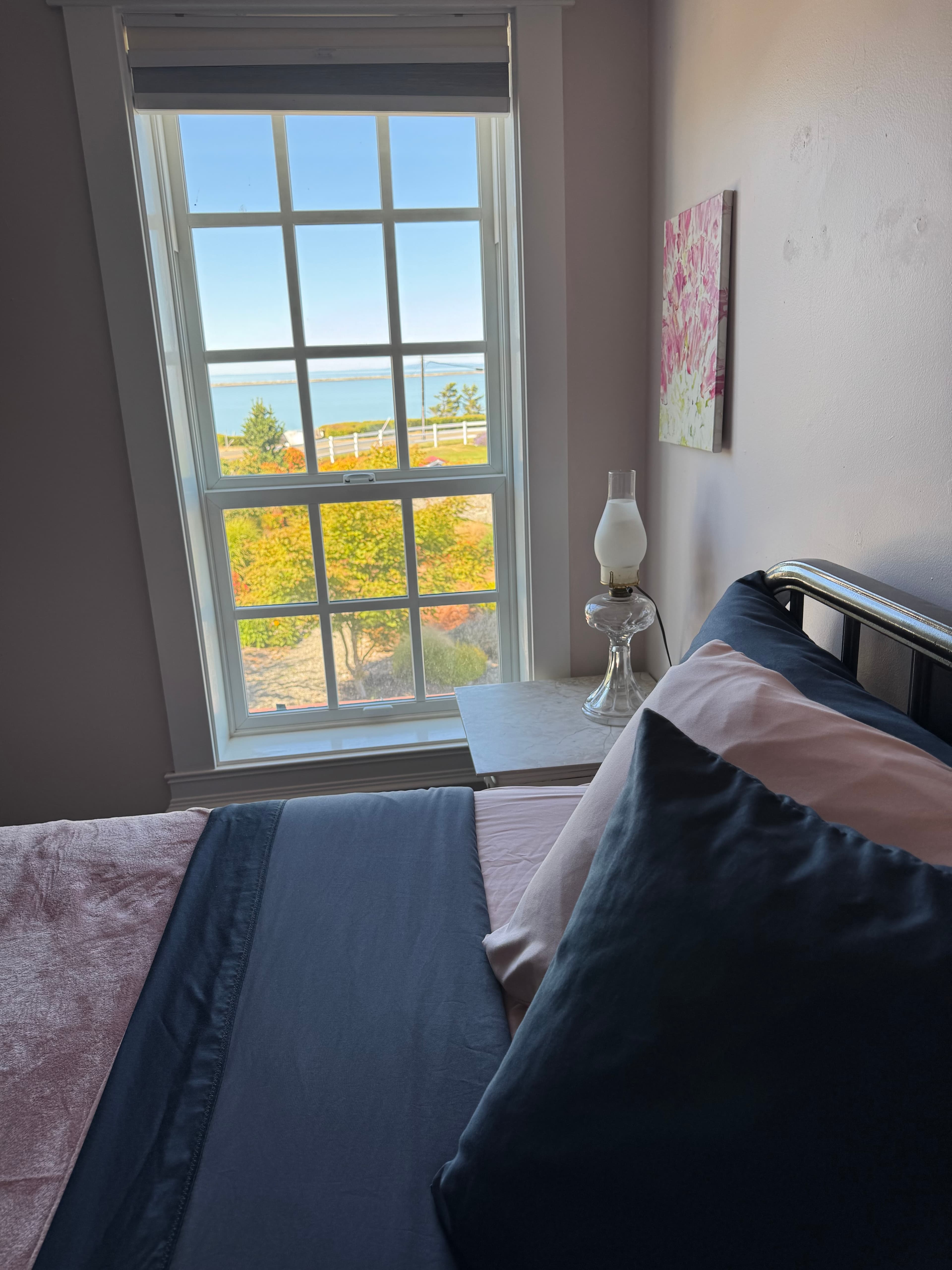 A cozy bedroom featuring a black metal bed frame with dark blue and pink linens, situated next to a large white-framed window that offers a clear view of the blue ocean and green trees beyond.
