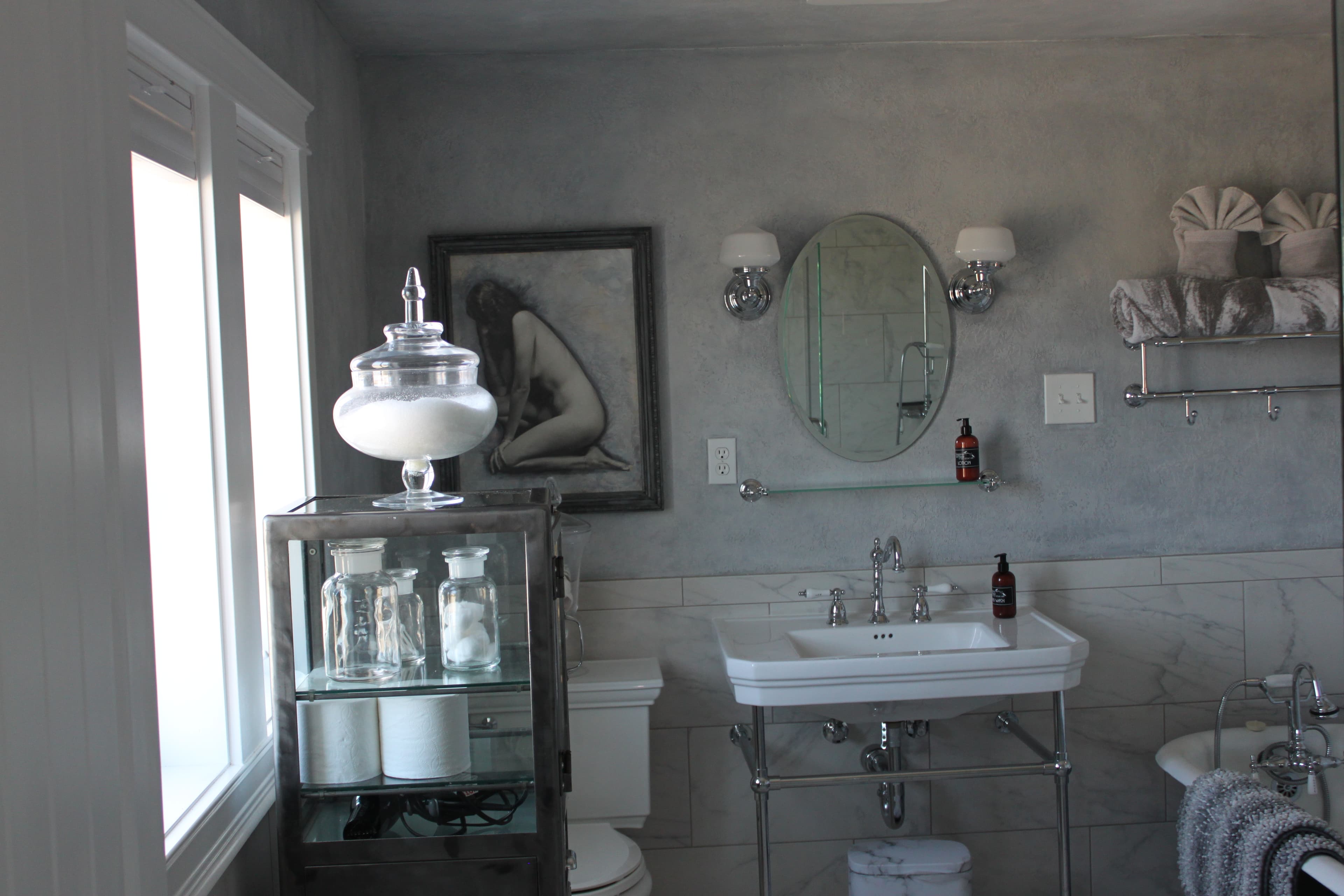 A spacious, well-lit bathroom with light grey walls and marble tiling, featuring a pedestal sink with an oval mirror, a vintage-style glass display cabinet filled with apothecary jars, and a classic clawfoot tub.