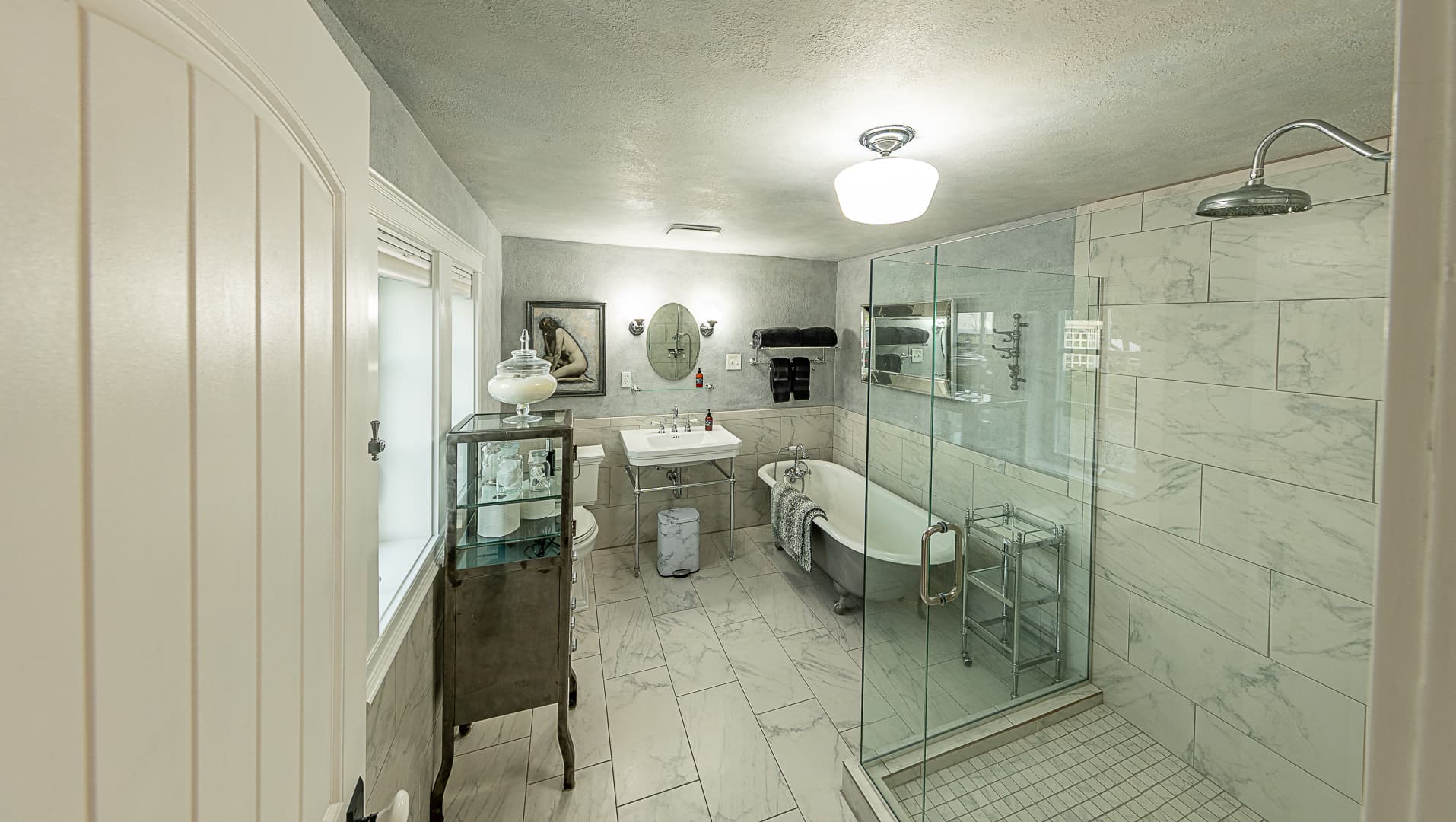 A spacious, luxury bathroom with white marble-style tiling, featuring a large glass-enclosed walk-in shower on the right and a classic clawfoot soaking tub in the center.