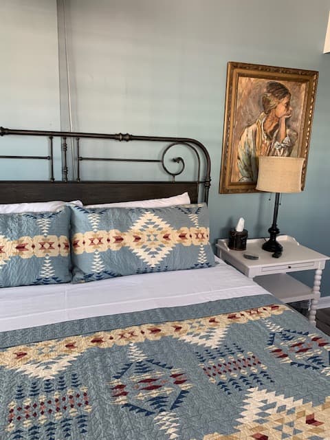 An elegant king-size bed featuring blue southwestern-patterned linens and a dark metal headboard, positioned next to a white nightstand with a classic lamp and an ornate framed portrait on a soft blue wall.