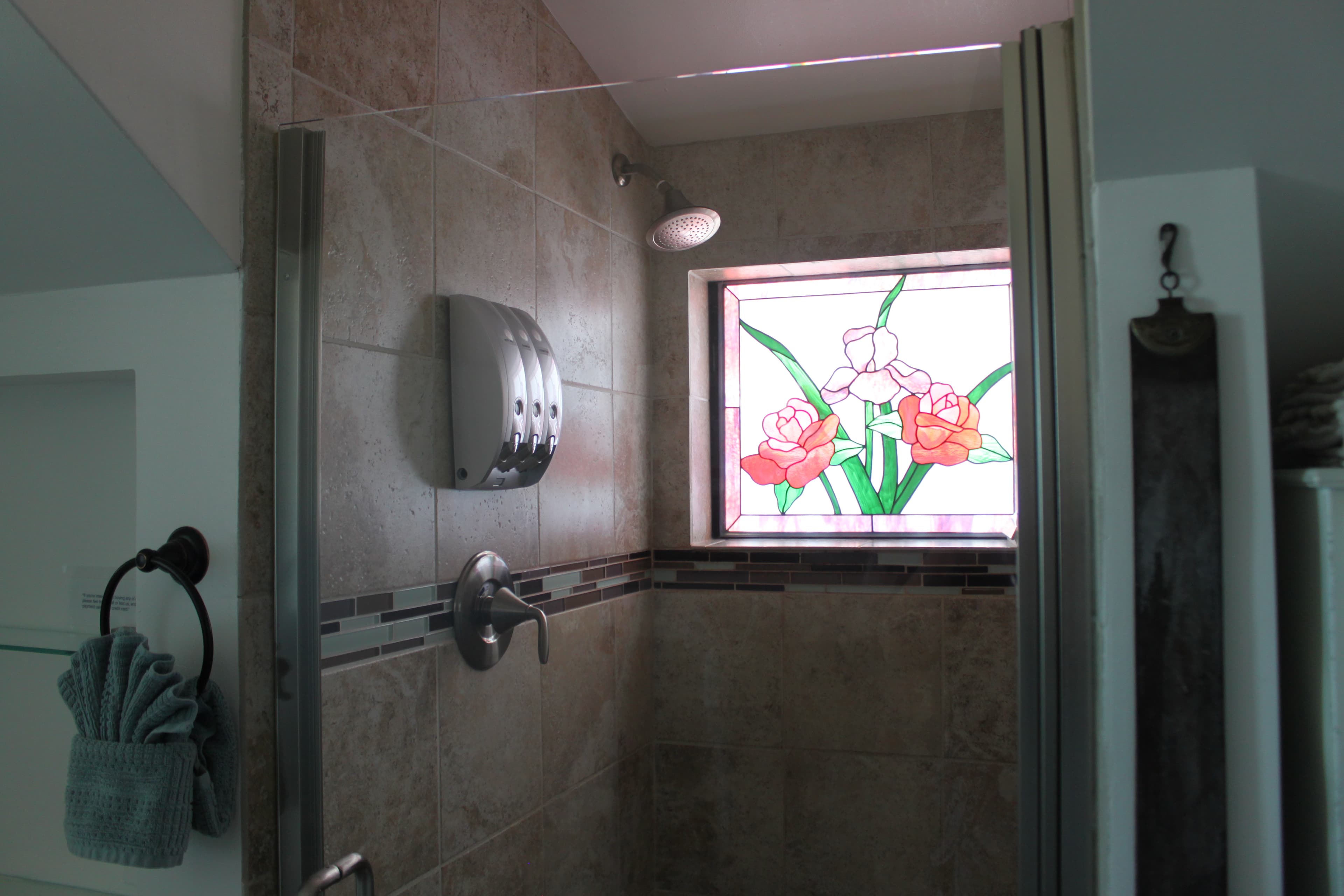 A close-up of a vibrant, square stained-glass window in a spacious, tiled, walk-in shower, featuring two red roses and a light pink iris with green leaves against a white background.