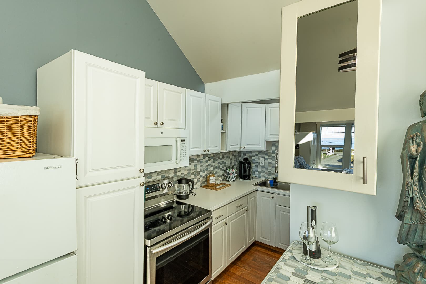 A close-up view of a modern kitchenette with white shaker-style cabinets, a stainless steel electric range, and a patterned tile backsplash, featuring a glass-door upper cabinet that reflects a bright window.