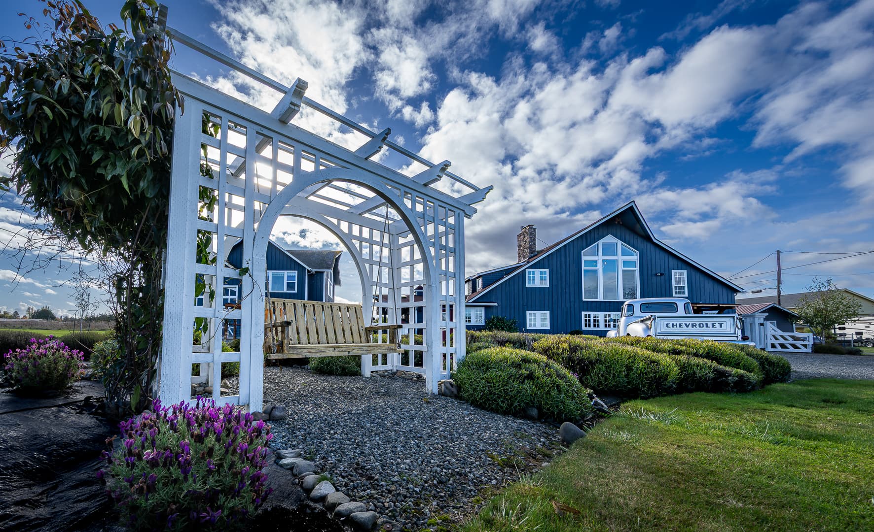 A white trellis with a swing in a garden setting, featuring a blue house and cloudy sky in the background.