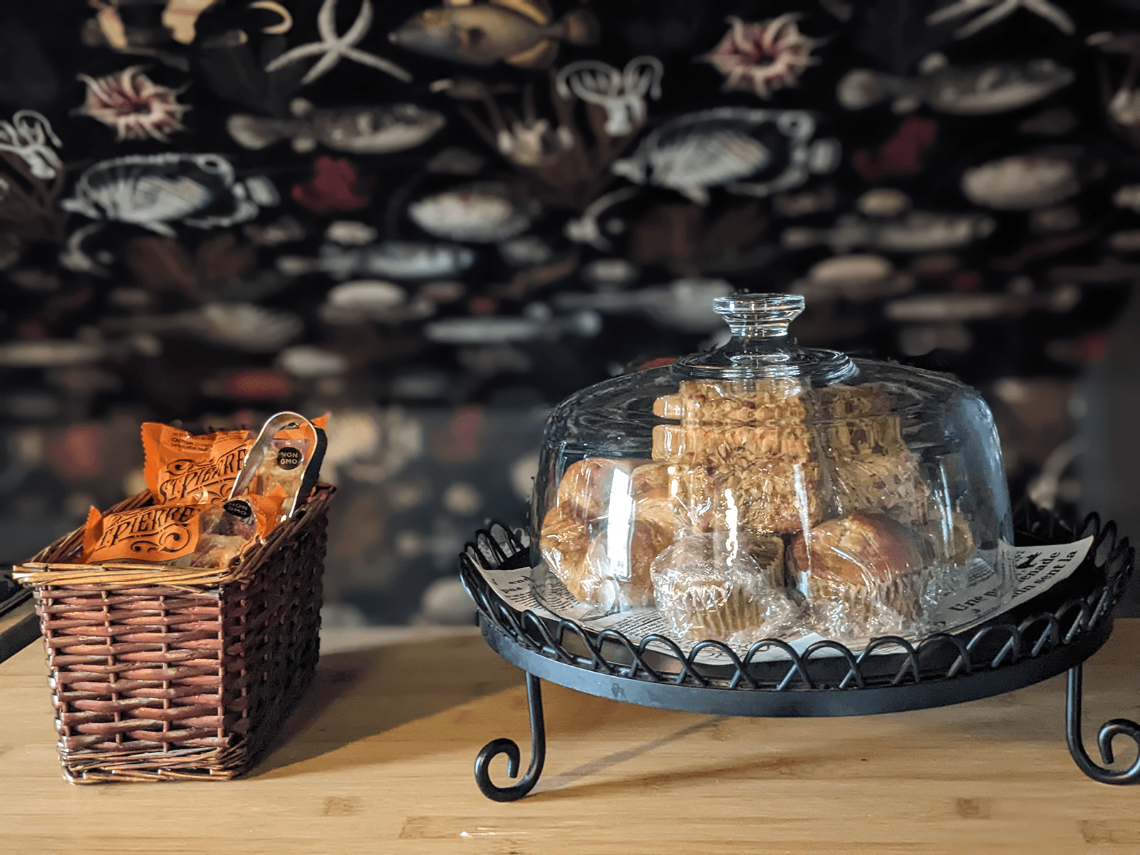 A glass cloche covers an assortment of pastries on a wire stand, next to a woven basket filled with snacks.