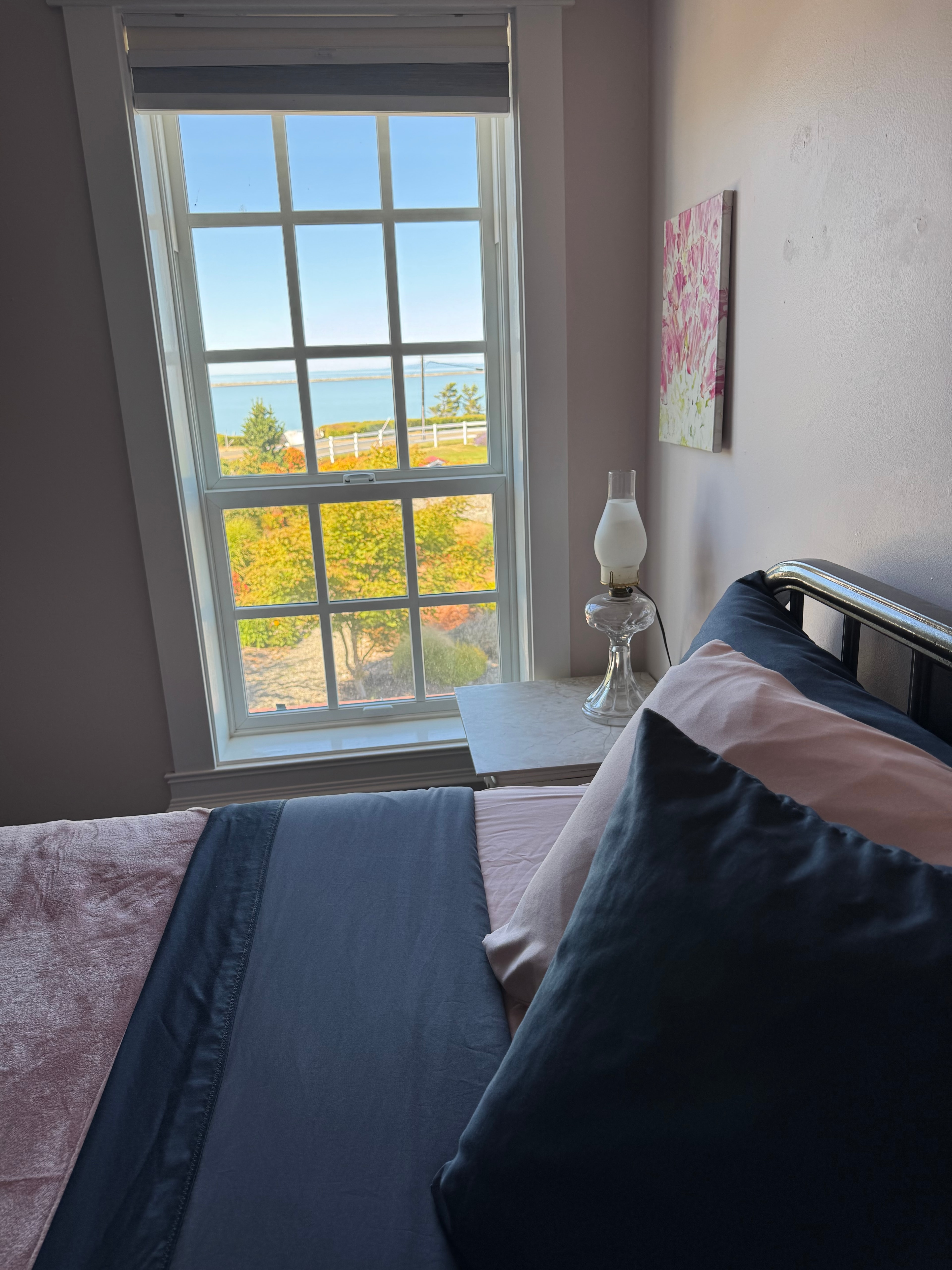 A cozy bedroom featuring a black metal bed frame with dark blue and pink linens, situated next to a large white-framed window that offers a clear view of the blue ocean and green trees beyond.