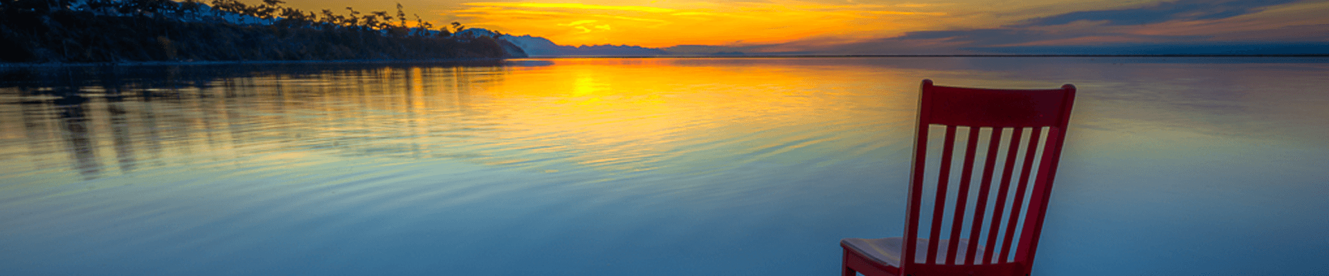 A red chair sits beside a calm lake at sunset.