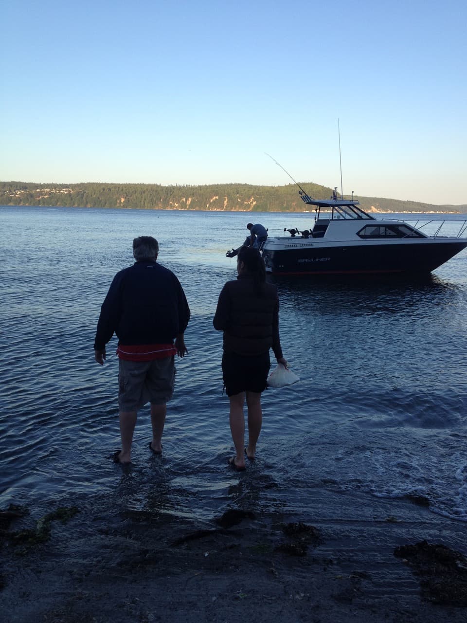 Two people stand in shallow water near a boat on a calm lake during sunset.