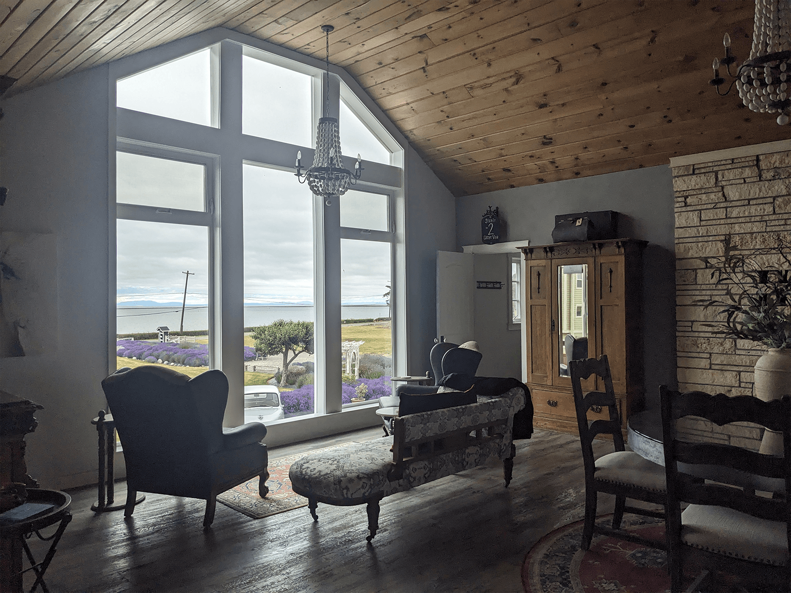 A cozy living room with large windows overlooking a scenic view, featuring vintage furniture and a wooden ceiling.