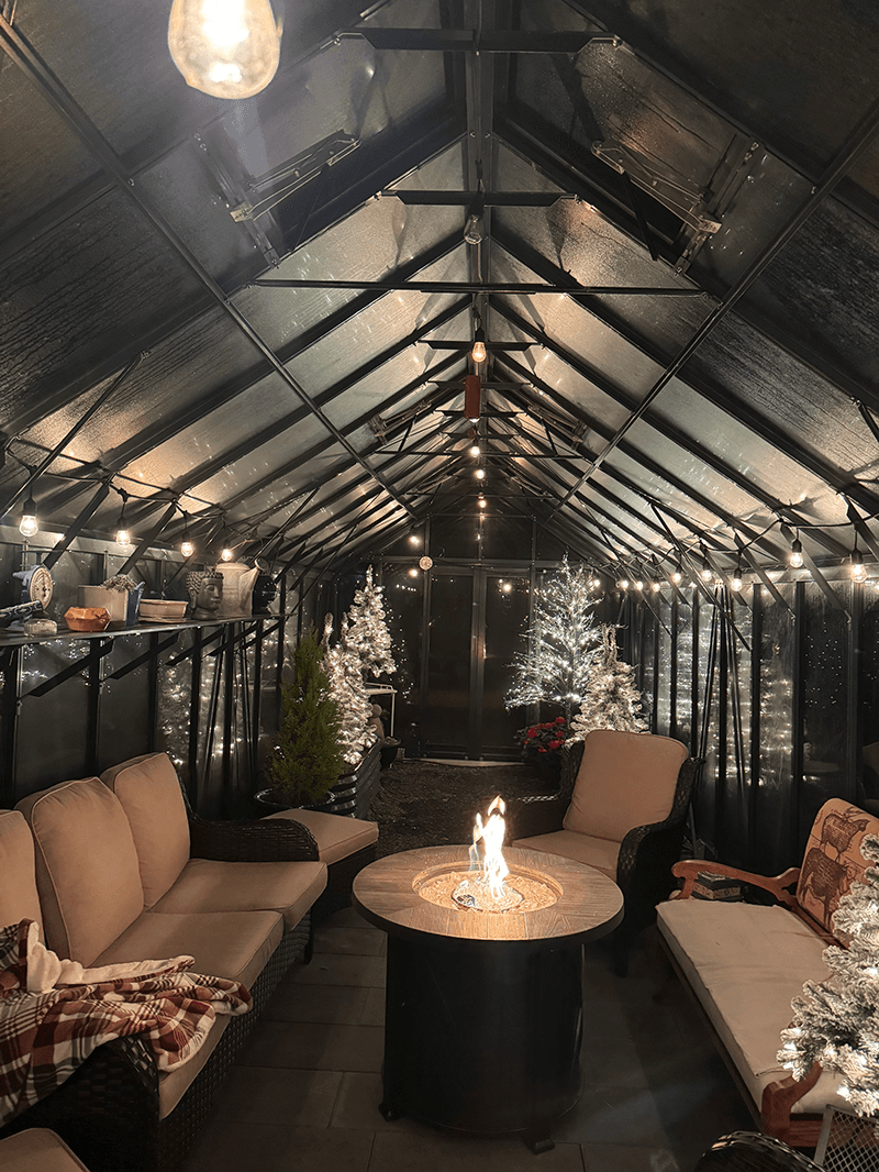 A cozy greenhouse interior decorated with twinkling lights and festive trees, centered around a fire pit and comfortable seating.